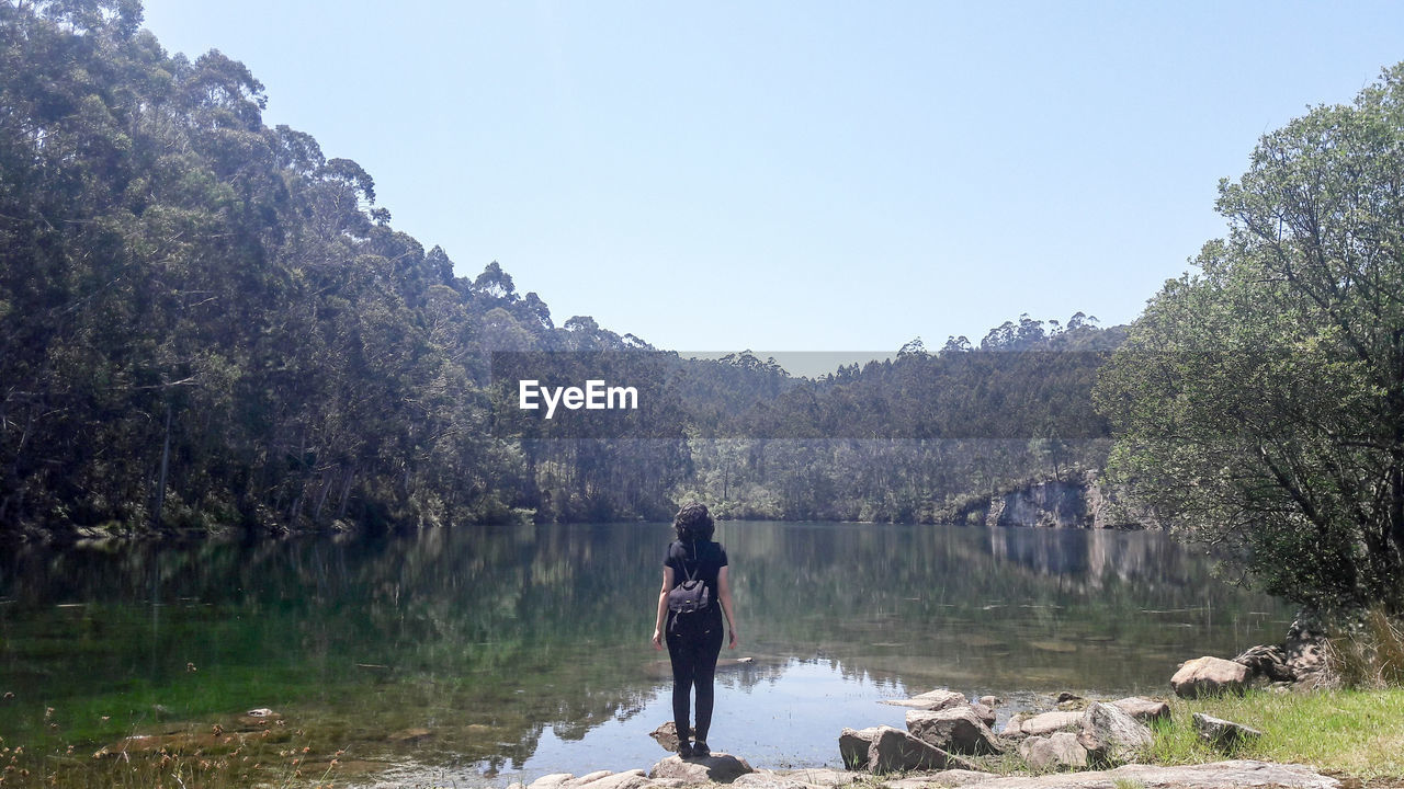 Rear view of woman with backpack standing by lake against clear sky