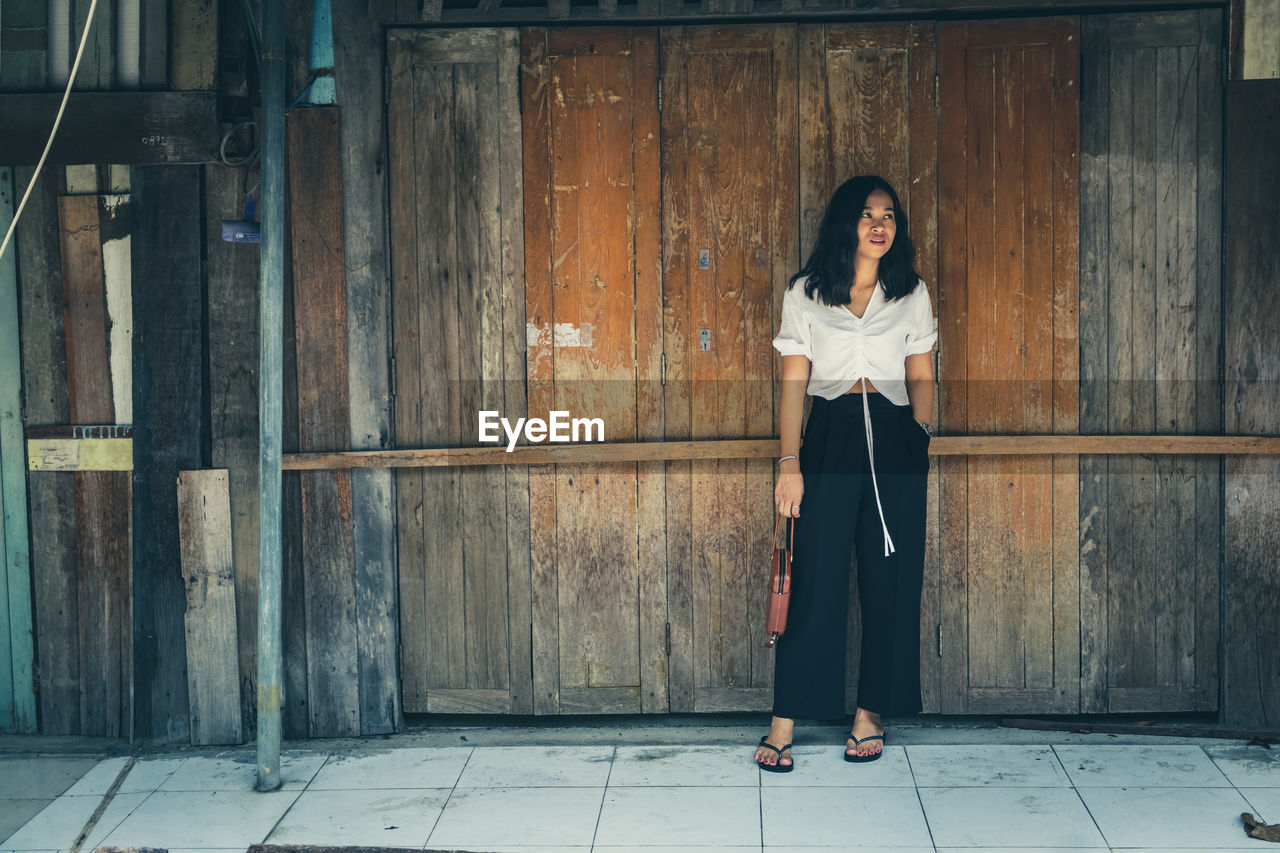 Full length of thoughtful woman standing against wooden wall