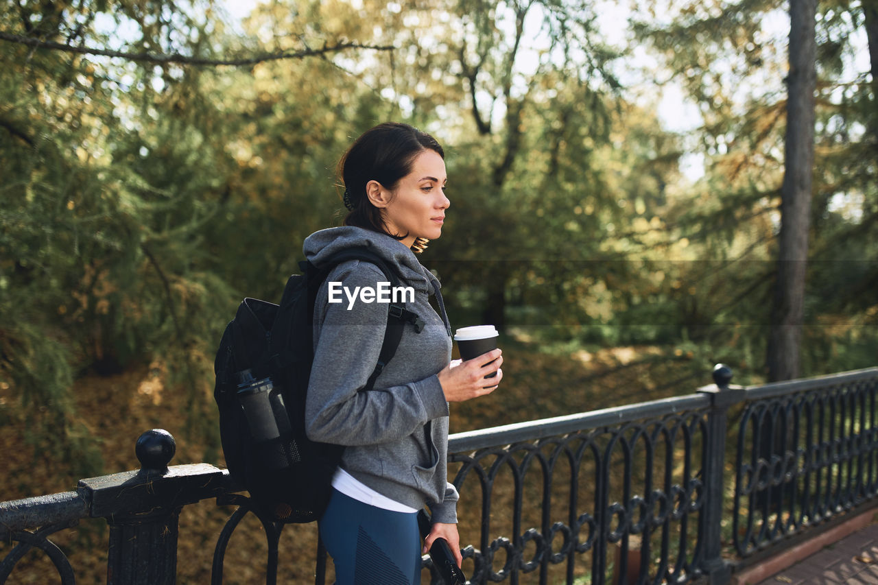 A brunette girl in a hoodie in nature walks in the park in autumn weather with coffe cup of paper