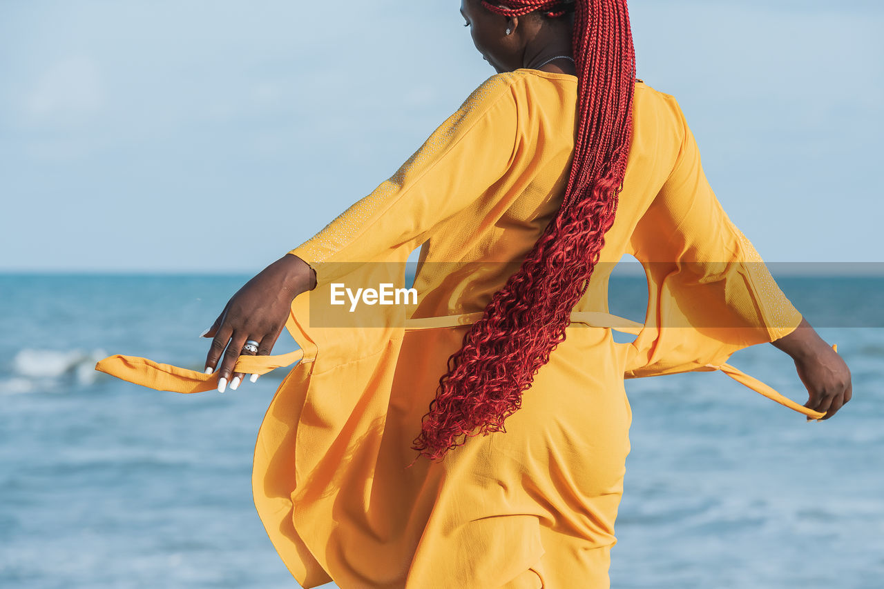 African woman wearing orange suit with long red hair standing by the sea at a sunset in accra 
