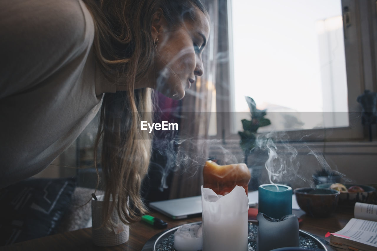 Side view of young woman blowing scented candles on table against window in dorm room