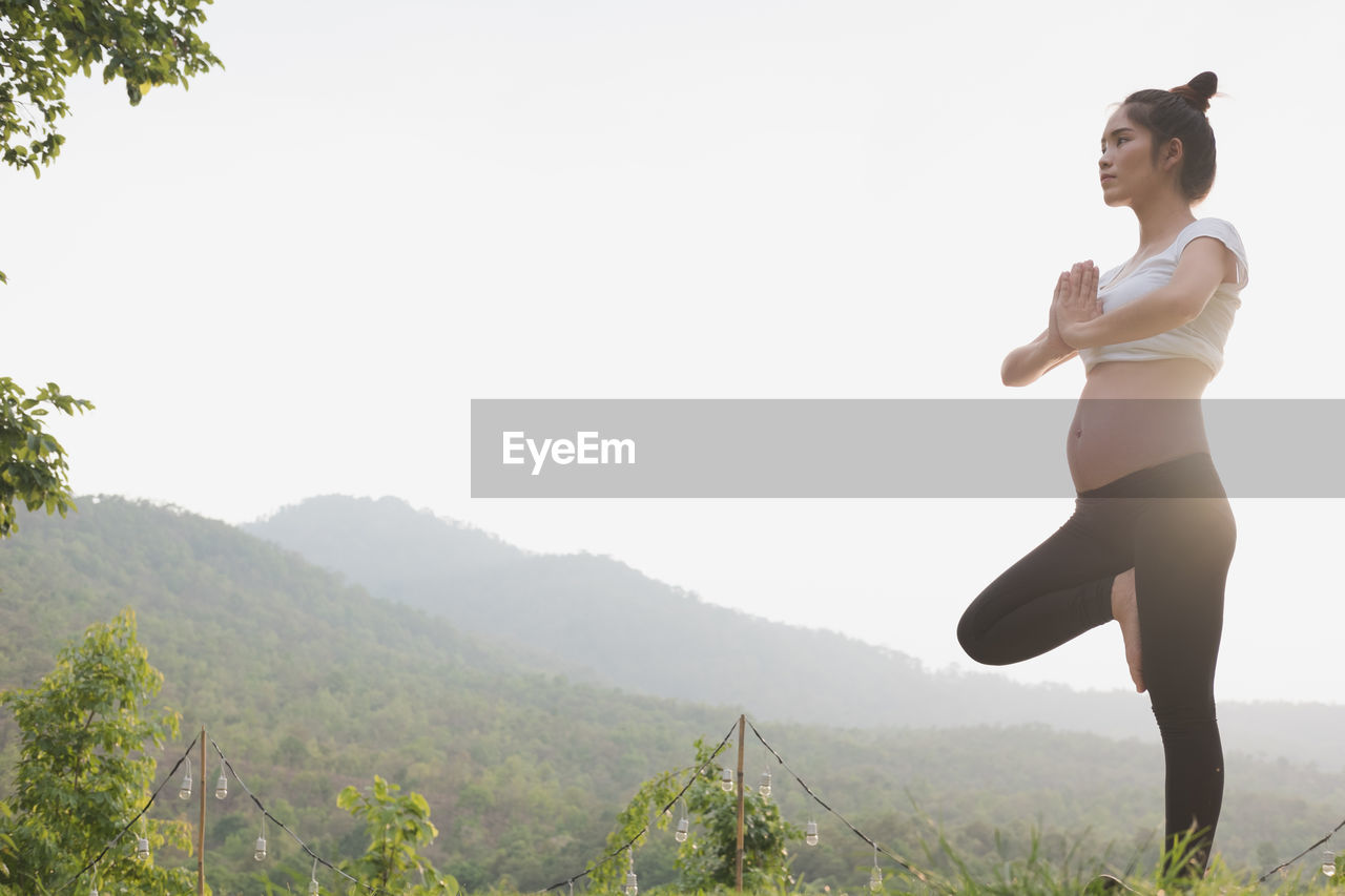 Pregnant woman exercising while standing on one leg against sky