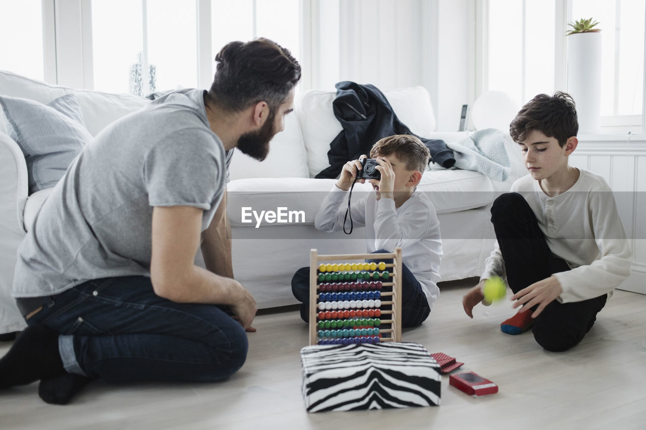 Boy photographing father by brother playing with ball in living room at home