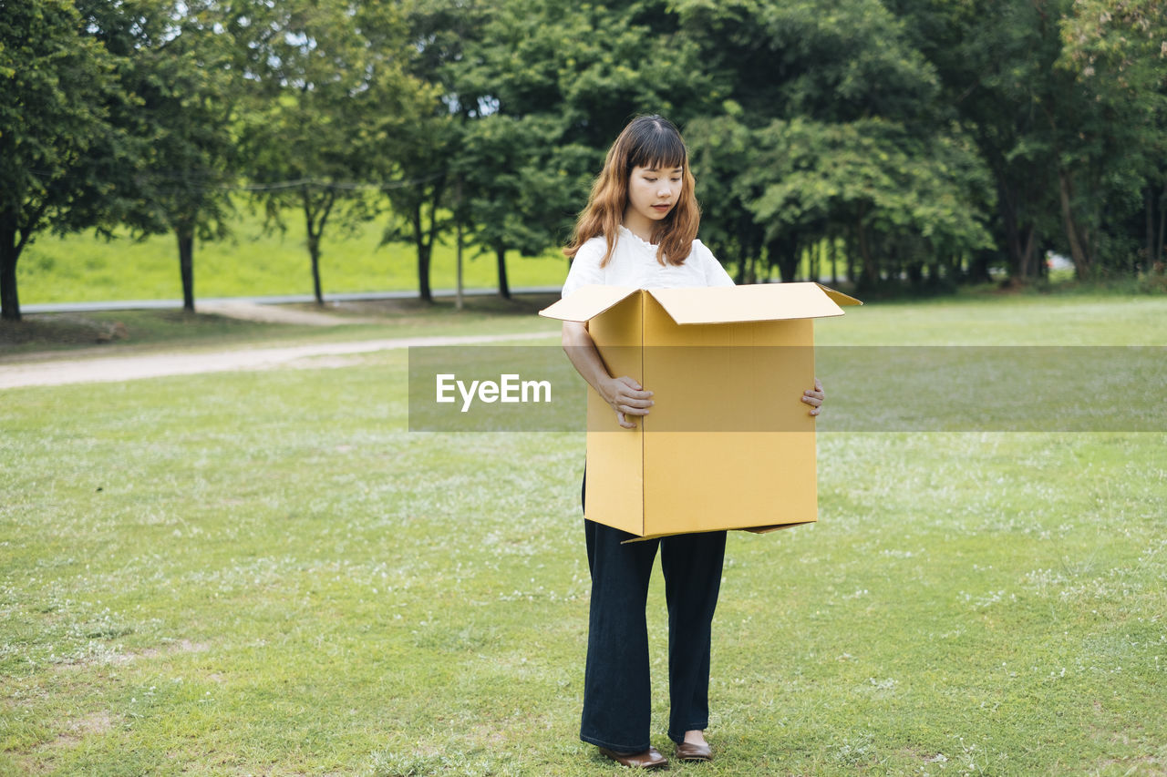 Full length of woman holding box while standing on grass against trees