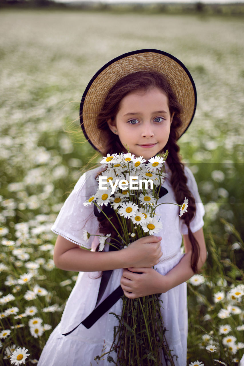 Portrait girl child in a white dress stands on a camomile field in a hat. bouquet of flowers