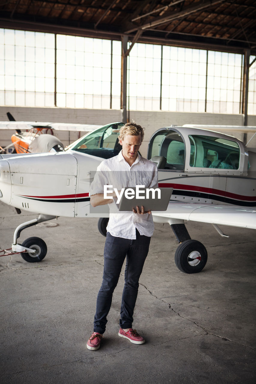 Man using laptop computer while standing by propeller airplane at hangar