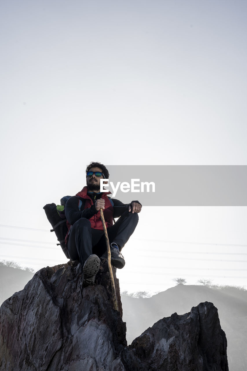 Young indian mountaineer sitting on the top of the mountain on a cliff with a backpack and a stick.