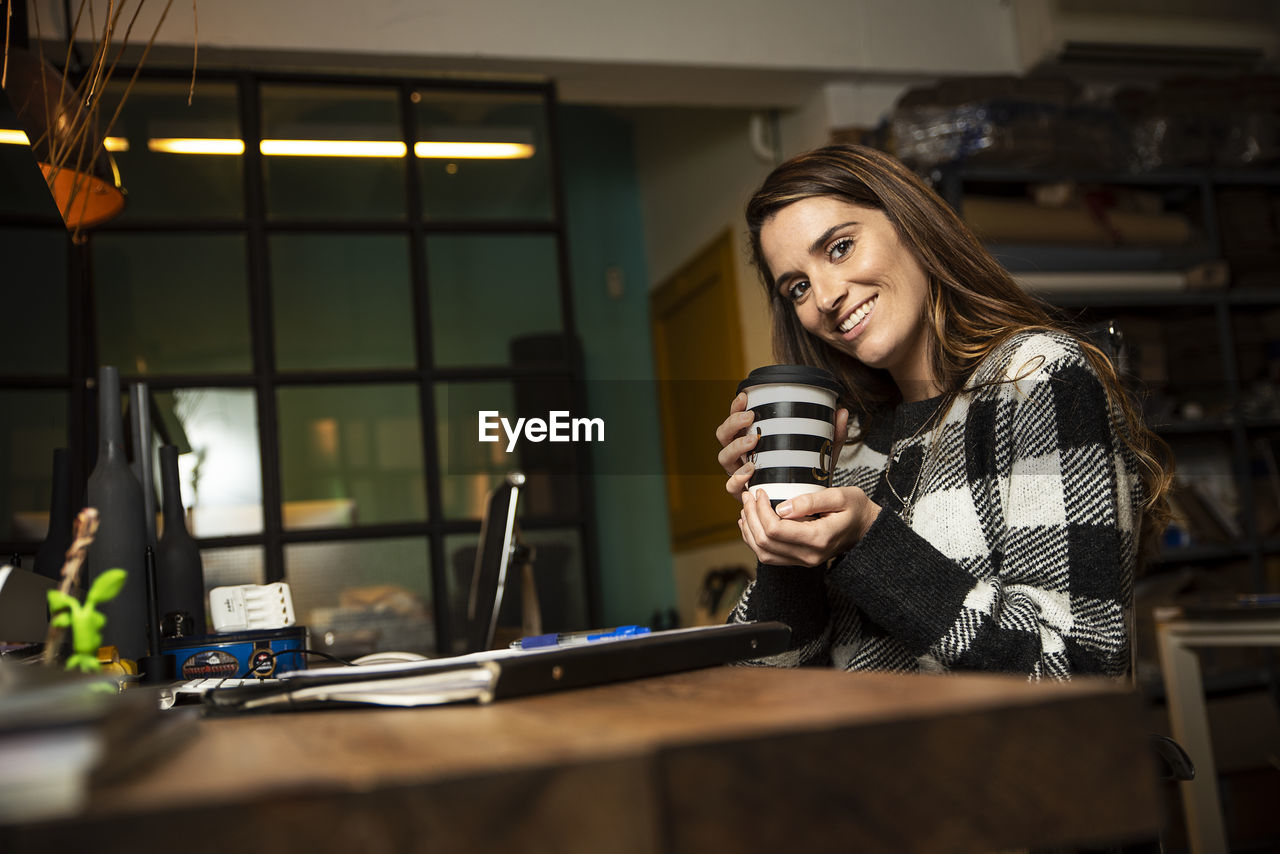 Portrait of smiling businesswoman holding disposable cup sitting at office