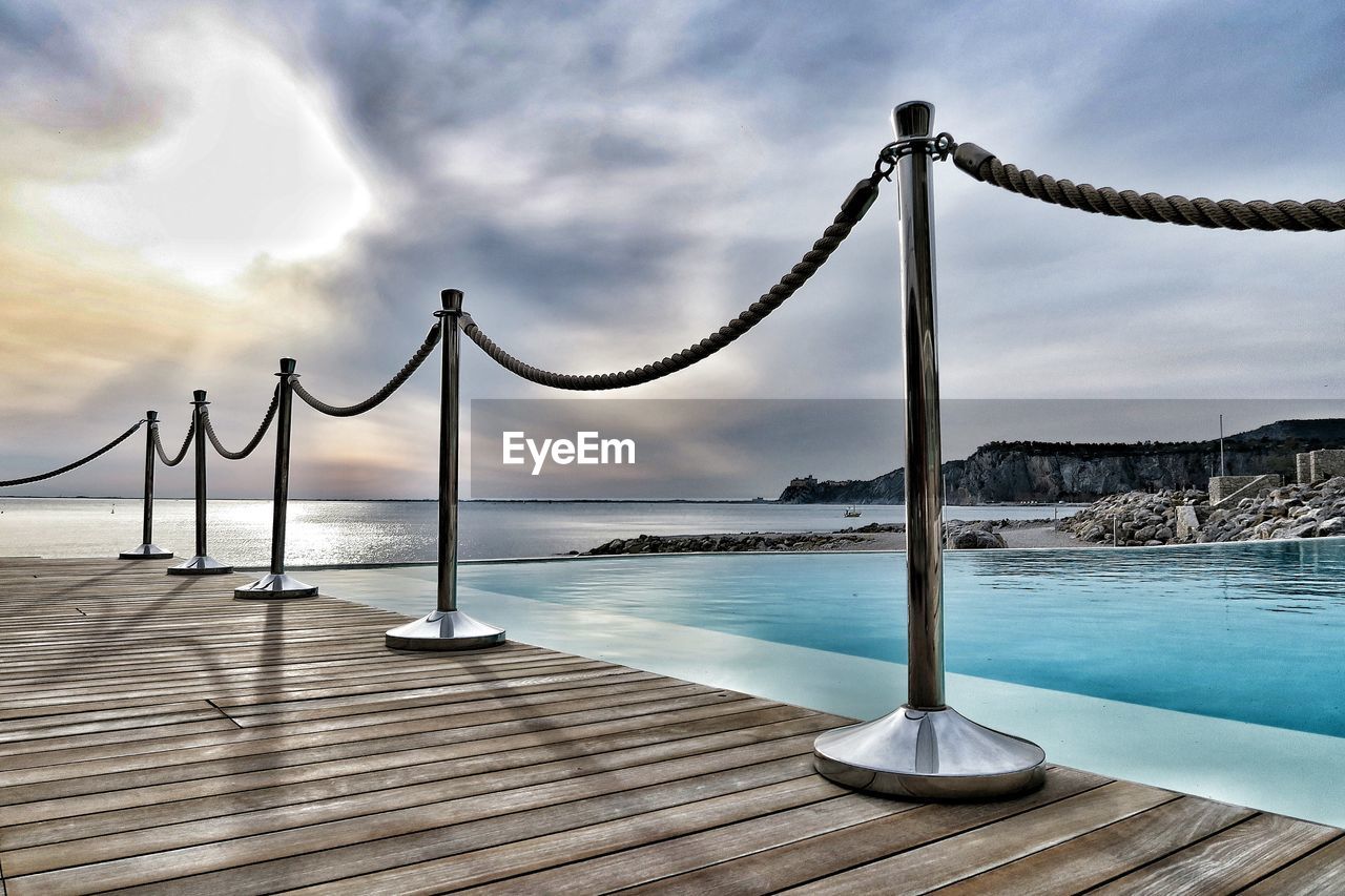 Railing on pier at poolside against cloudy sky