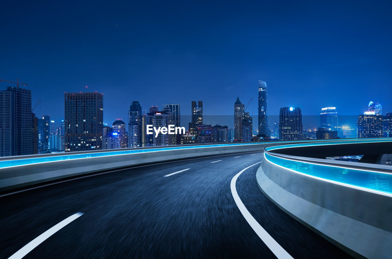 Light trails on road by illuminated buildings against blue sky at night