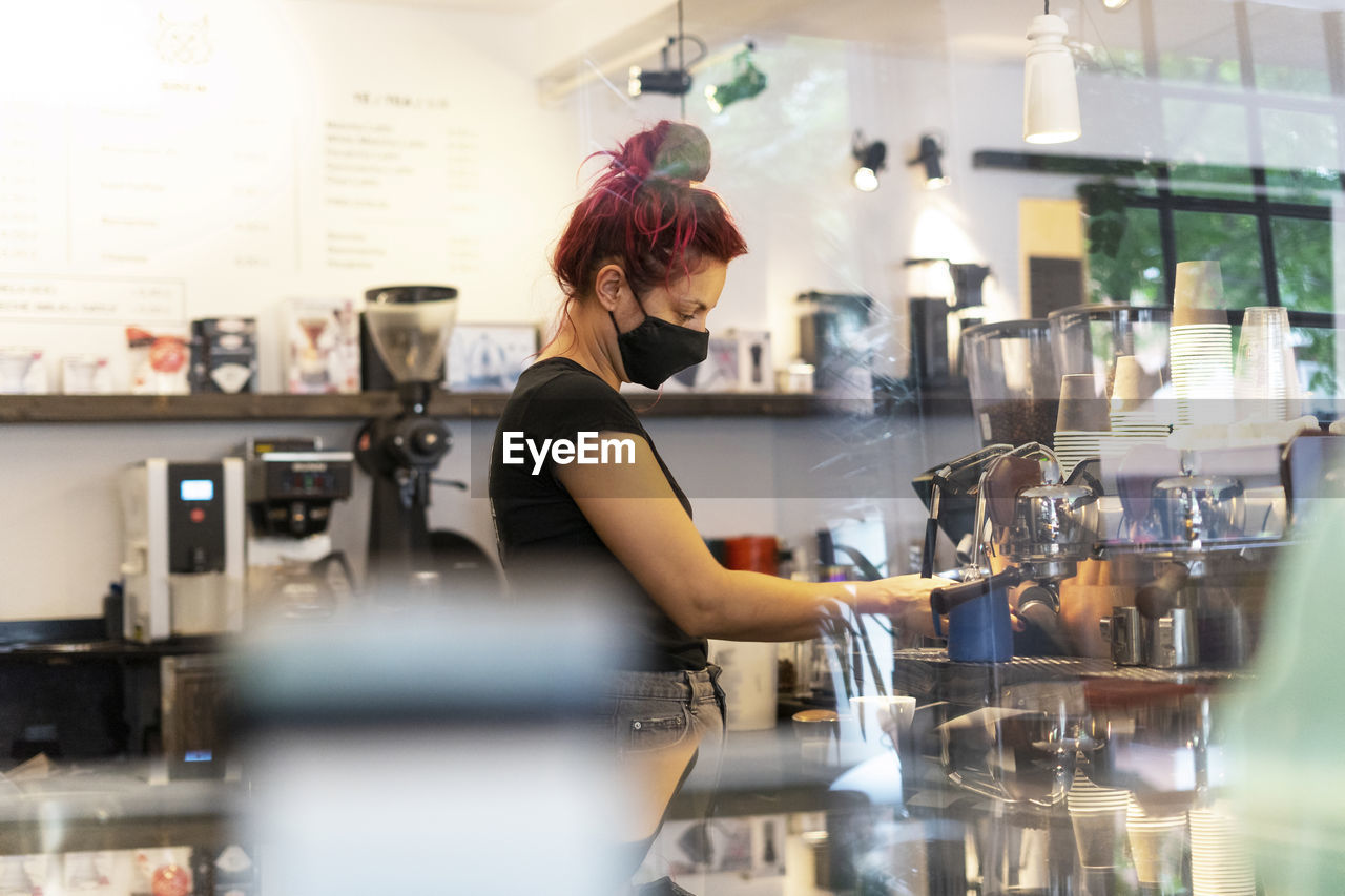 Side view of female bartender in face mask standing at counter in coffee shop and making delicious hot beverage in coffee machine during coronavirus epidemic