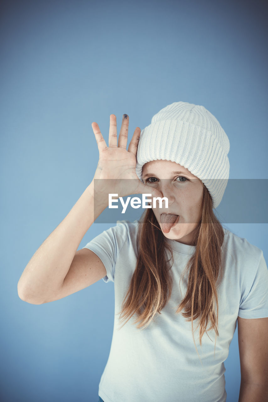 one person, cap, adult, portrait, studio shot, women, blue, clothing, colored background, finger, blue background, indoors, waist up, emotion, young adult, hand, person, white, looking at camera, gesturing, headgear, female, casual clothing, human face, front view, hat, hand raised, humor, fashion accessory, arm, hairstyle, standing, facial expression, long hair, photo shoot, fun, skin, human head, human limb, happiness, negative emotion, portrait photography, lifestyles, blond hair, smiling