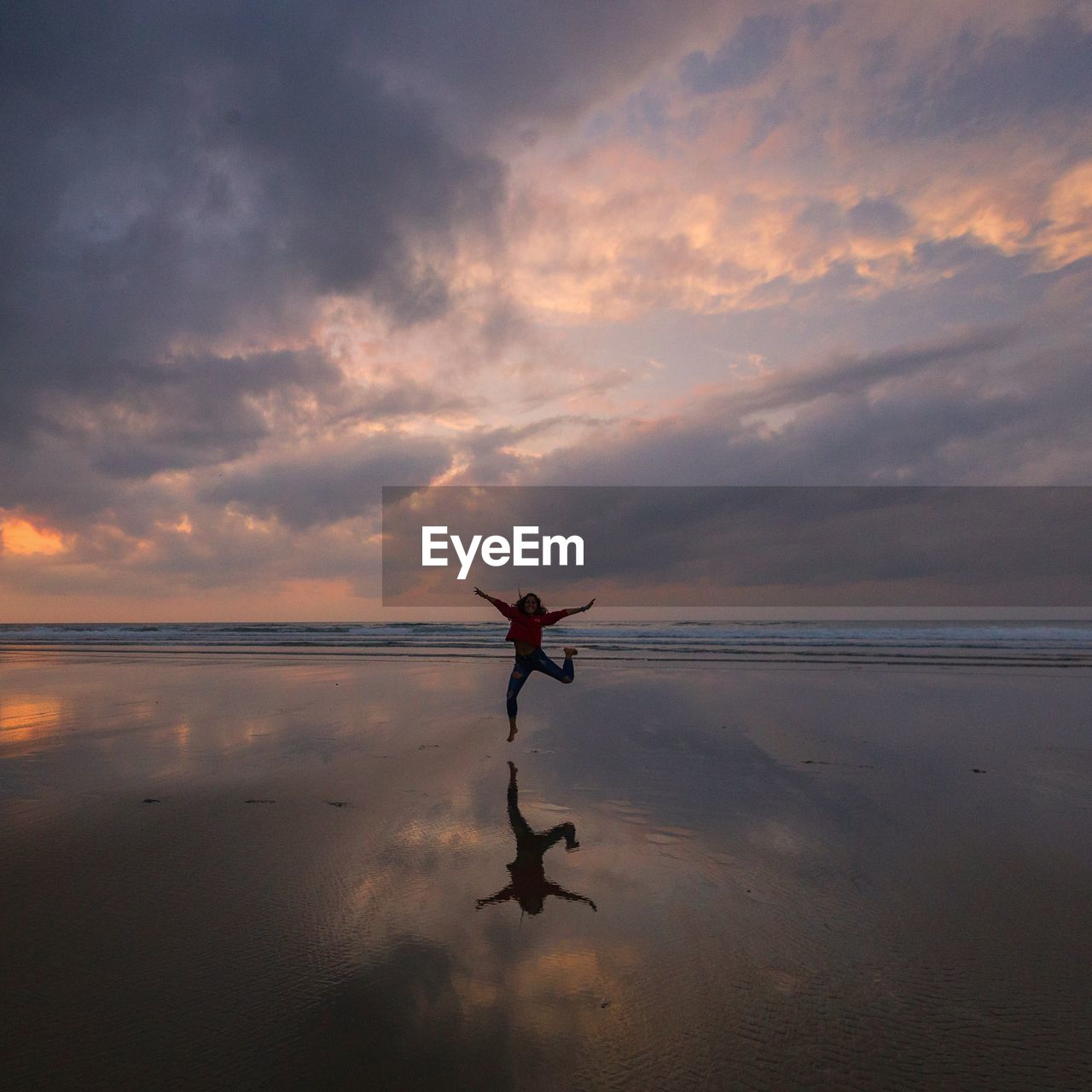 Woman jumping at beach against cloudy sky during sunset