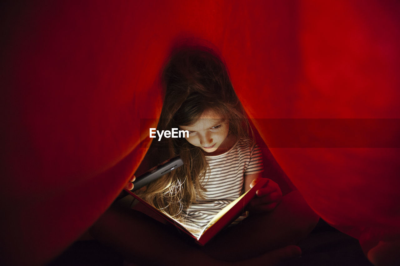 Close-up of girl with mobile phone light reading book under blanket