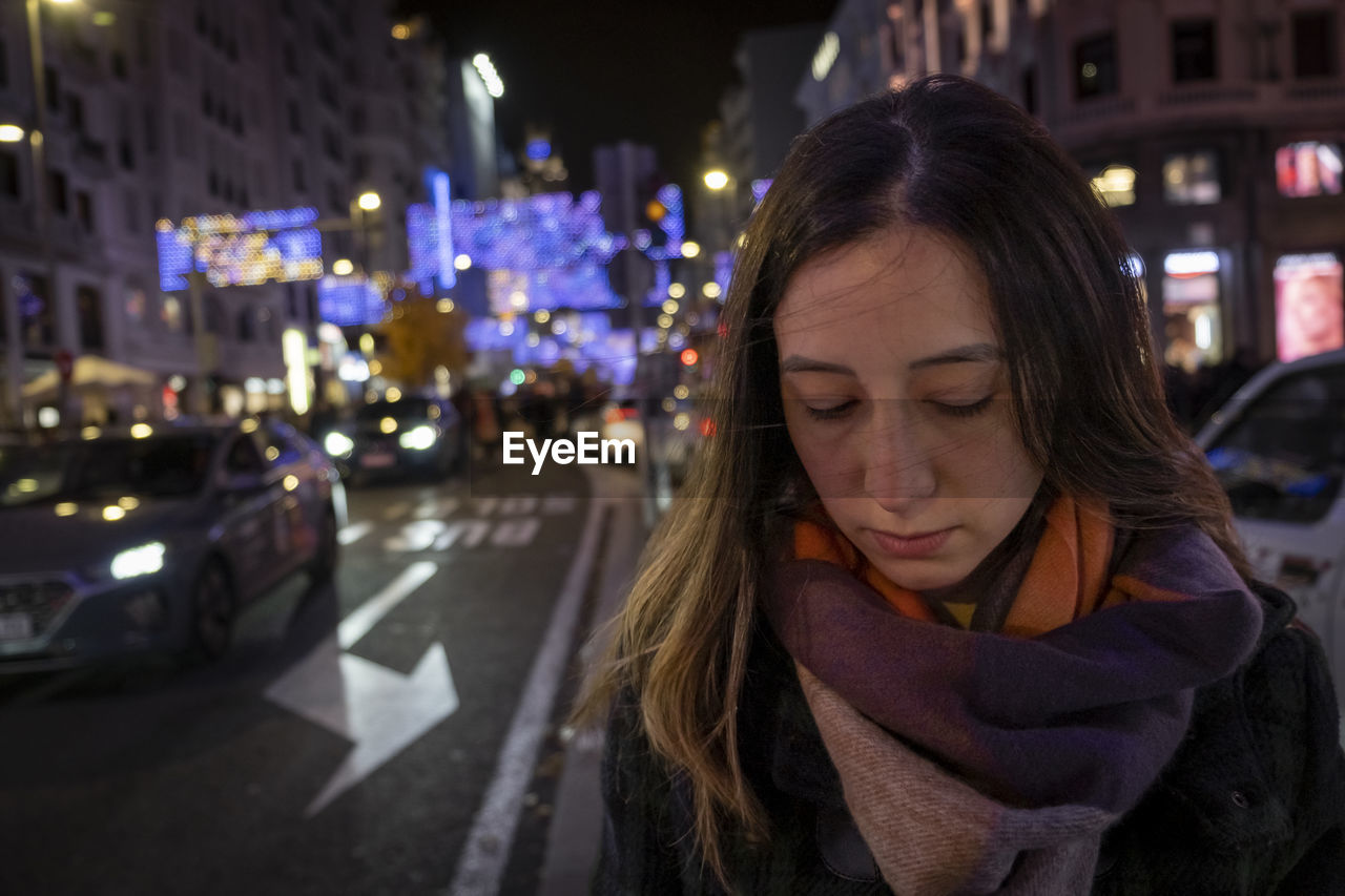 city, night, one person, street, city life, transportation, architecture, adult, women, illuminated, car, motor vehicle, city street, mode of transportation, headshot, looking down, portrait, focus on foreground, vehicle, young adult, looking, front view, scarf, lifestyles, long hair, standing, winter, evening, road, building exterior, person, brown hair, outdoors, hairstyle, waist up, human face