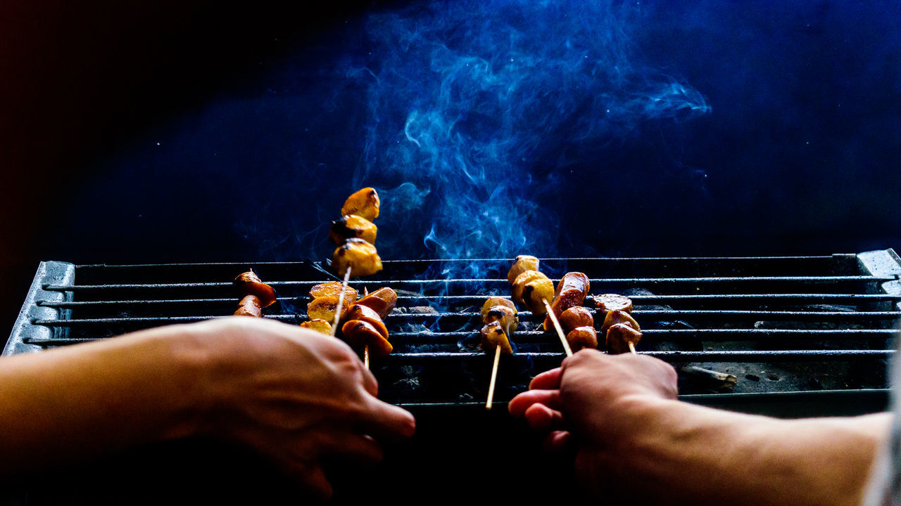 Cropped hands holding skewers with meat on barbecue grill at night