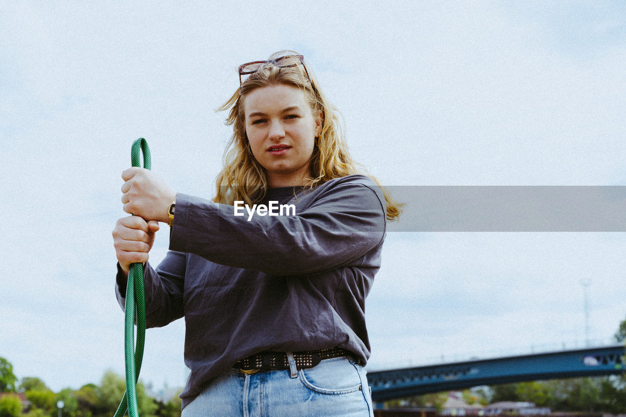 Low angle portrait of confident young blond woman holding garden hose against sky