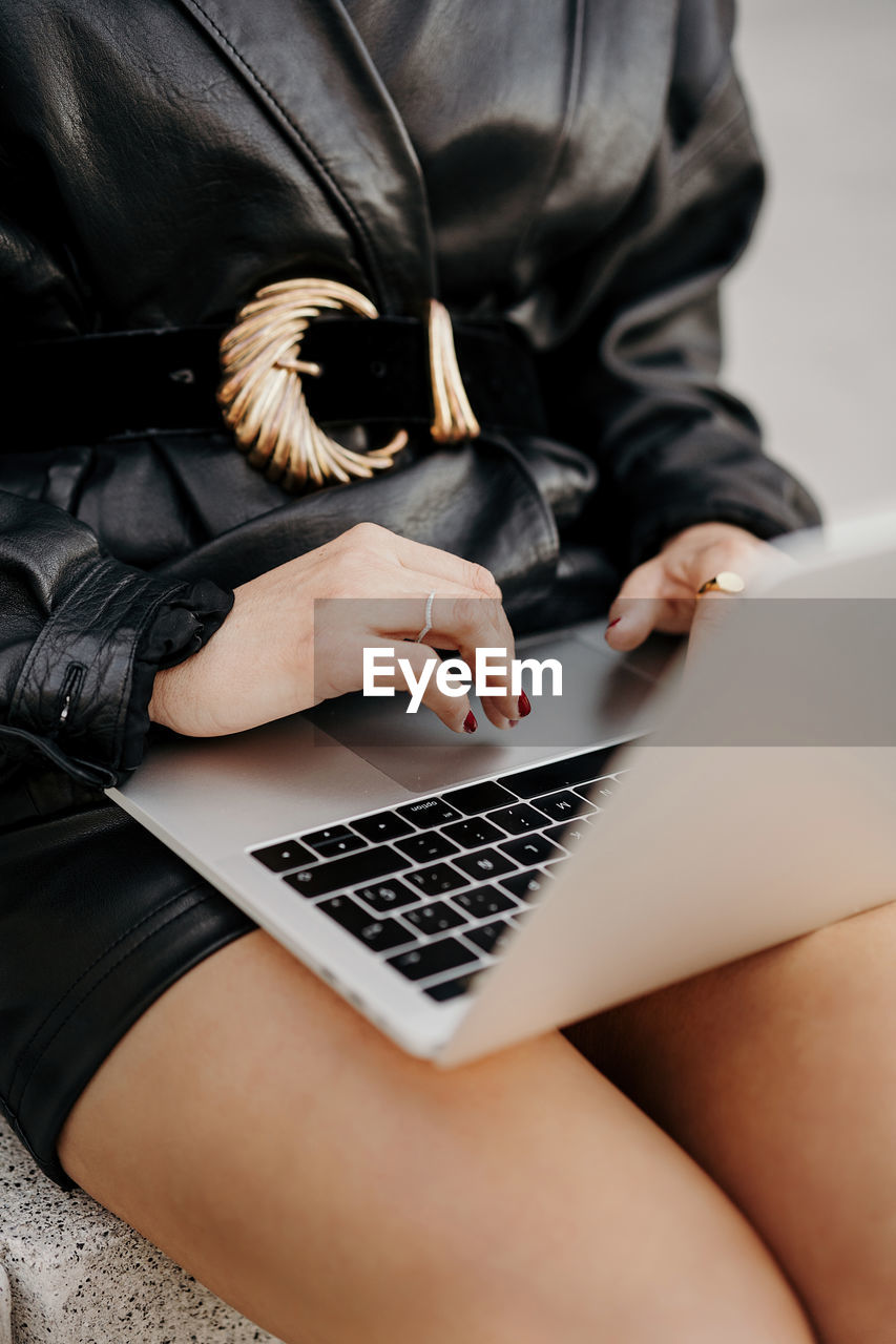 Clever curly businesswoman wearing black leather suit and jacket typing on laptop keyboard while sitting on stairs and working on remote project on city street