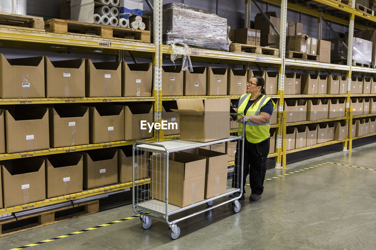 Full length of female worker pushing cart with cardboard boxes by rack at warehouse