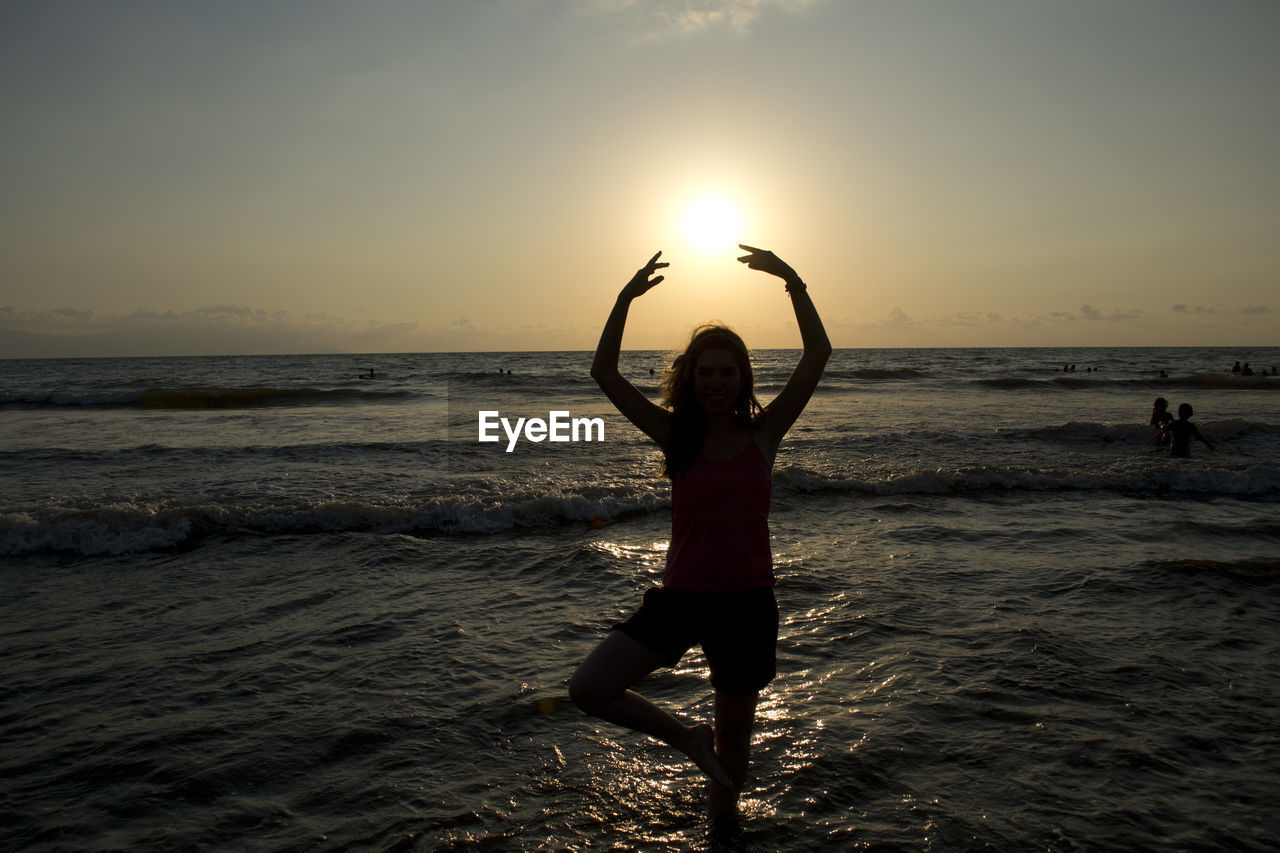 WOMAN STANDING ON BEACH AGAINST SEA DURING SUNSET