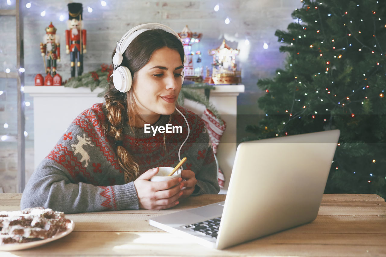 Woman with cup of coffee using laptop and headphones at christmas time