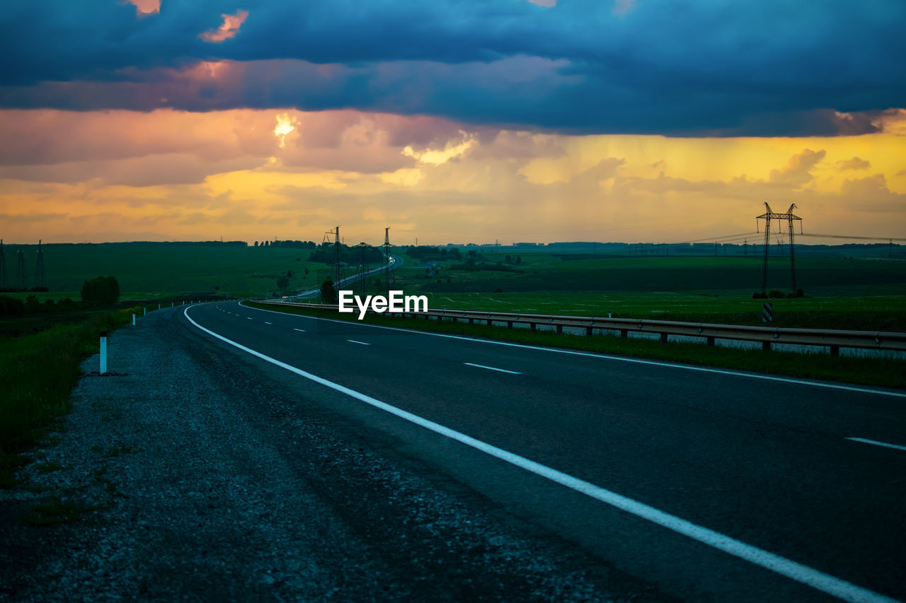 sky, cloud, road, transportation, horizon, environment, nature, sunset, landscape, dusk, dramatic sky, highway, beauty in nature, no people, street, scenics - nature, travel, city, sign, symbol, land, outdoors, evening, mode of transportation, the way forward, vanishing point, cloudscape, tranquility, marking, road marking, rural scene, moody sky, grass, sunlight, diminishing perspective, tranquil scene, travel destinations, field, architecture, horizon over land, asphalt, country road