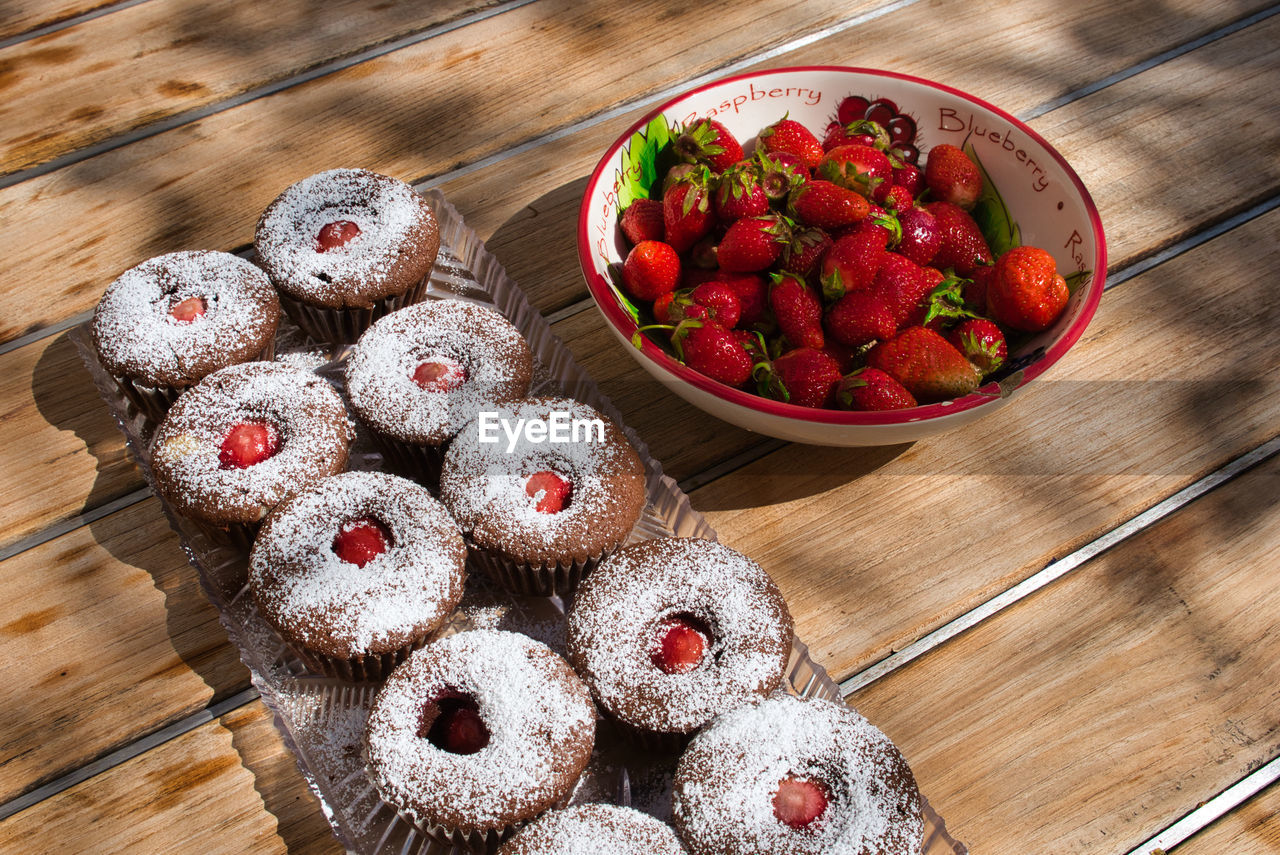 Homemade cupcakes next to bowl of ripe strawberries on wooden outdoor table. high angle view.