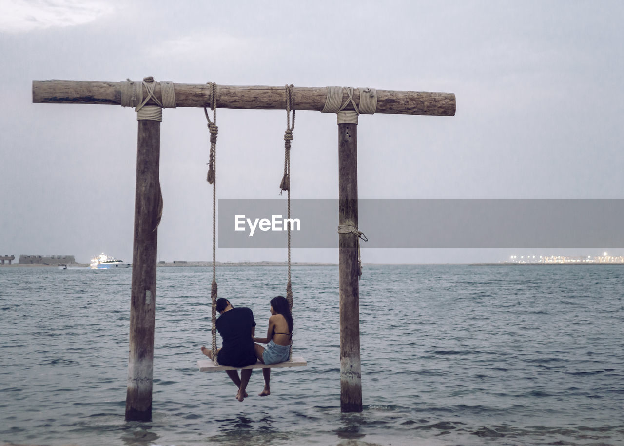 Rear view of couple sitting on rope swing over sea against sky