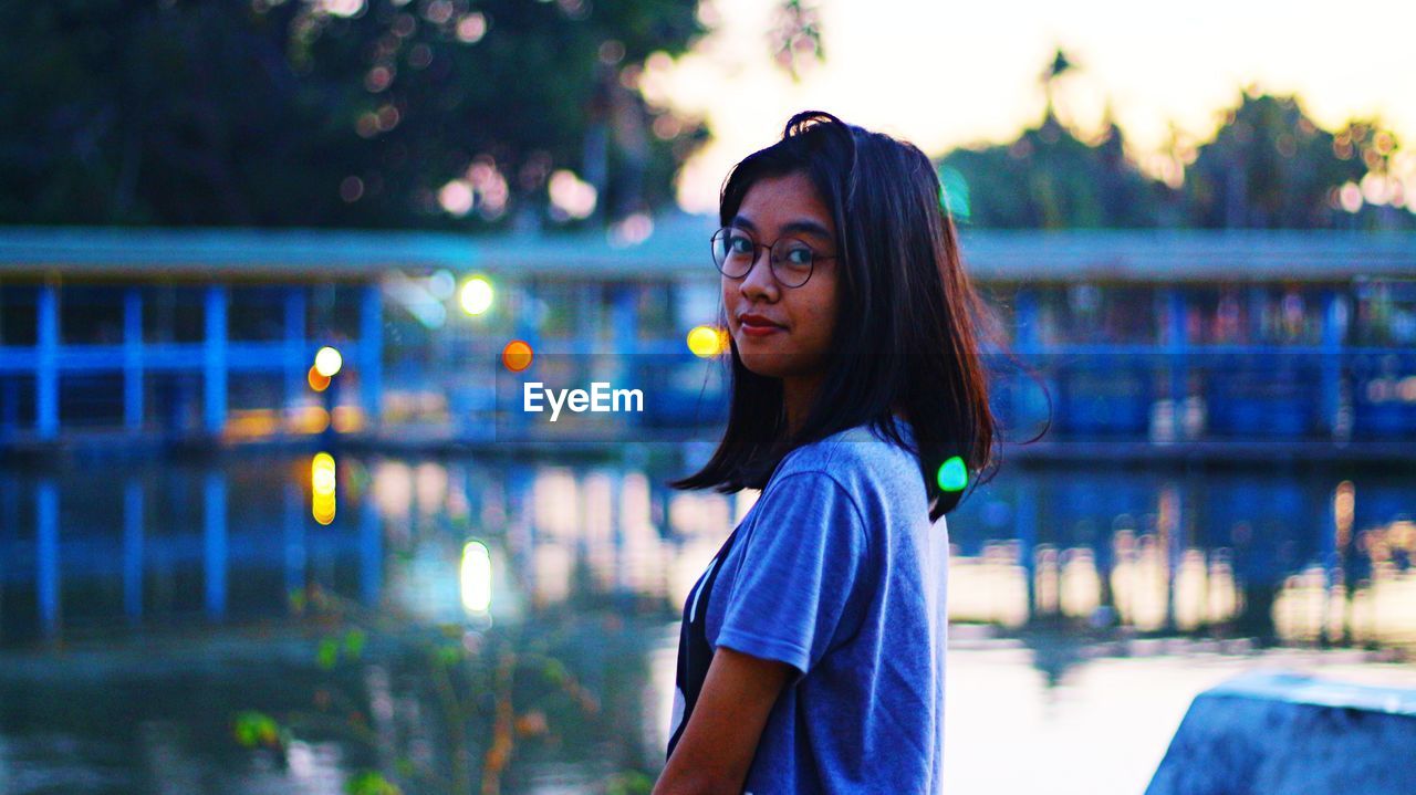 Portrait of young woman standing by lake during sunset