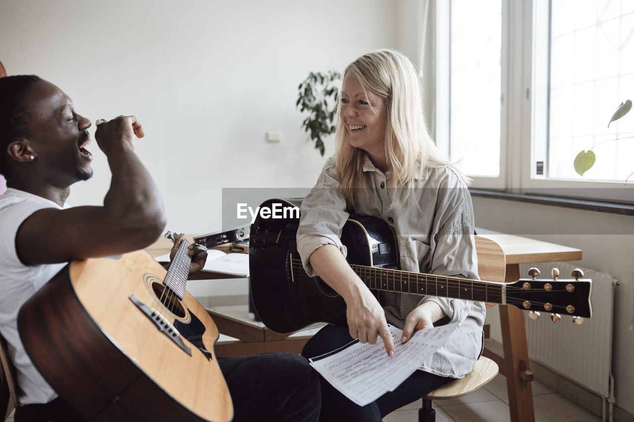 Happy female tutor looking at male student laughing while practicing guitar in classroom