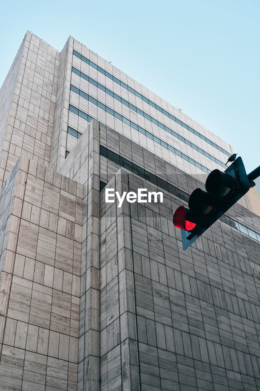 From below of black traffic light with red color and modern building with creative architecture under blue sky on background