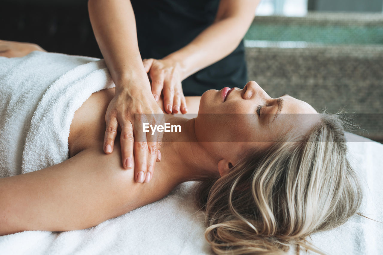 Attractive blonde young woman on couch with white linen enjoys hand facial massage in spa salon