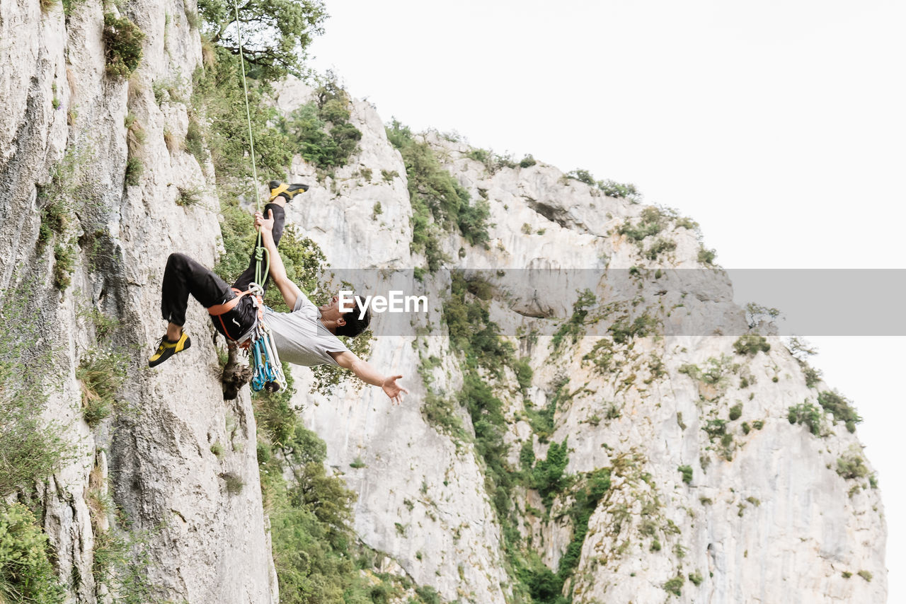 Full length of cheerful male hiker shouting while rappelling on rocky cliff against sky