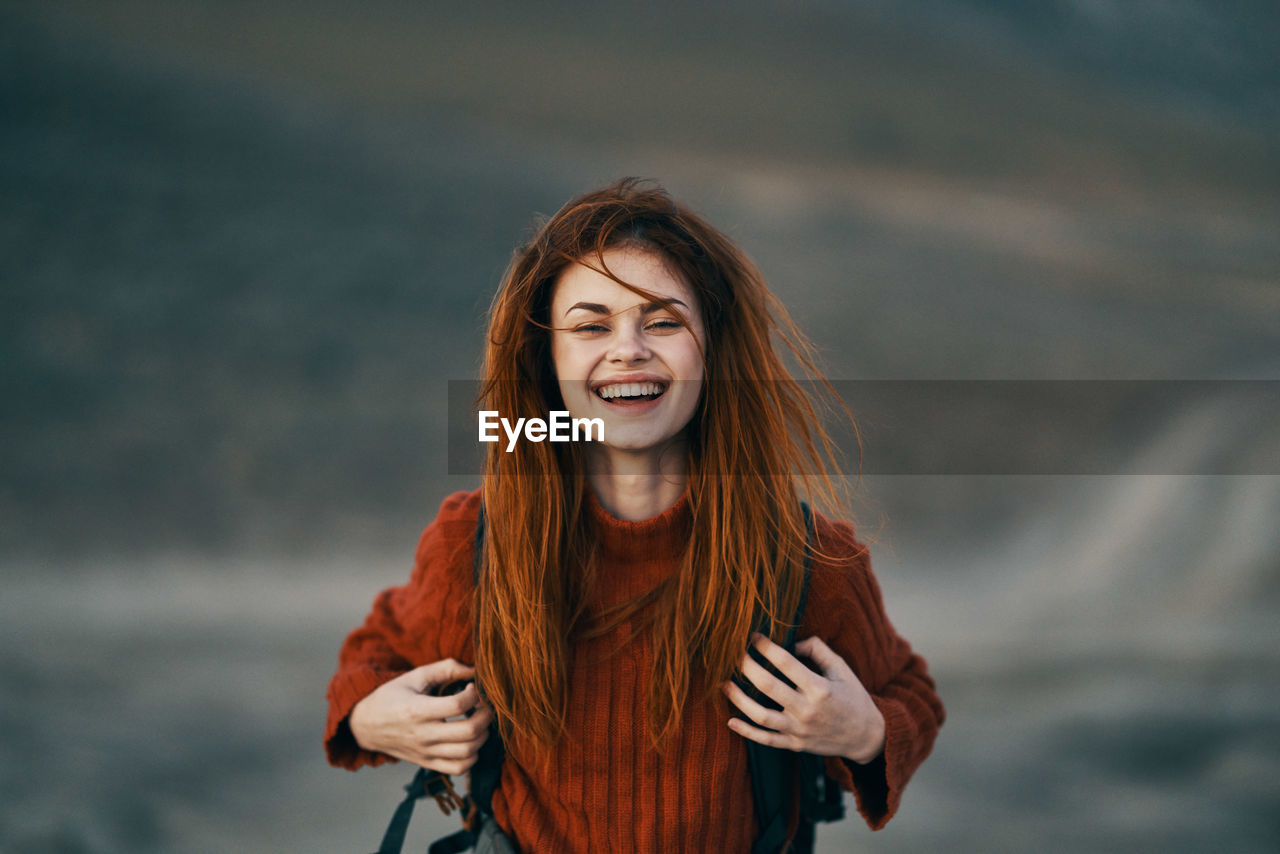 PORTRAIT OF SMILING YOUNG WOMAN STANDING AGAINST BLURRED BACKGROUND