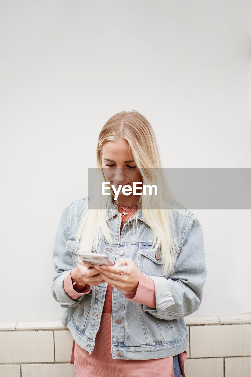 Young blonde woman looking on her smartphone whilst standing in front of a wall