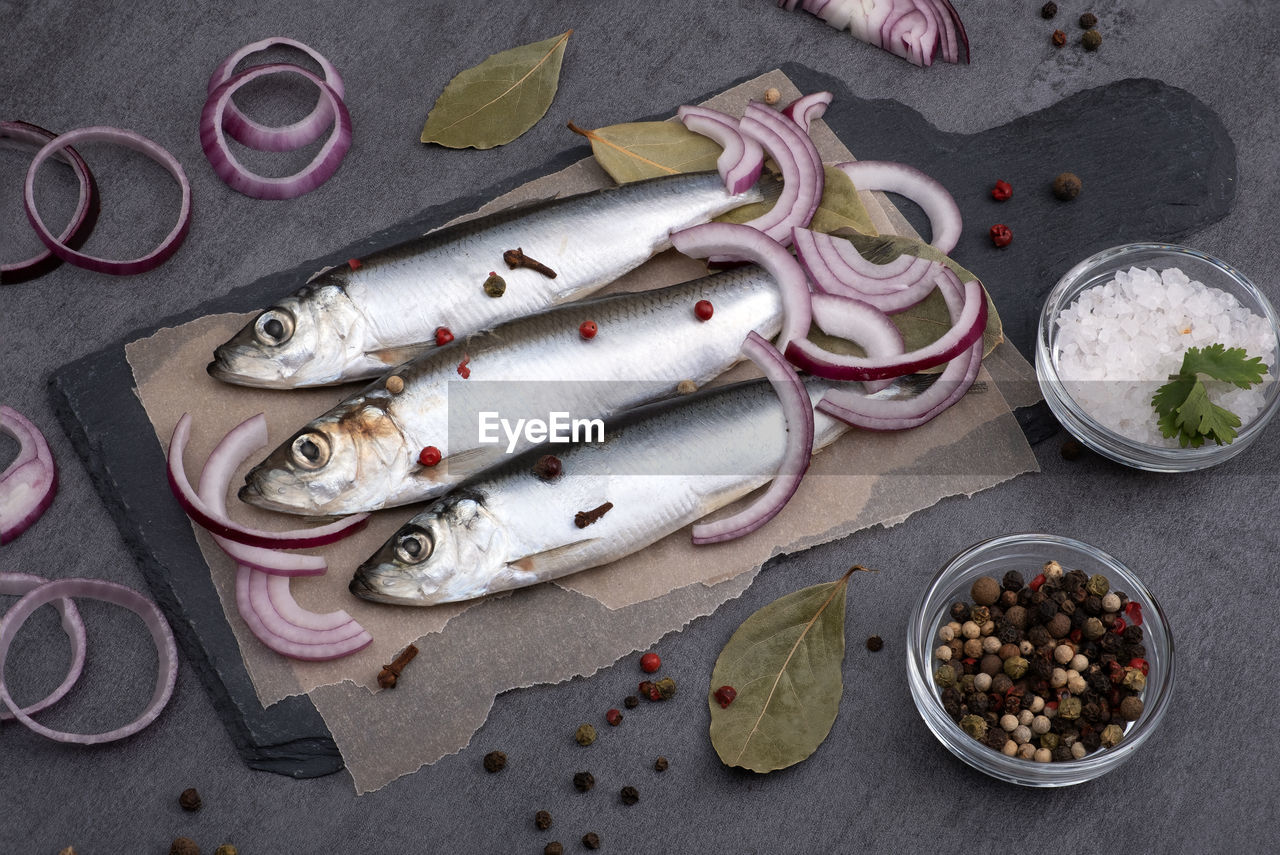 Close-up top view of several pickled herrings with spices on parchment paper on a serving board. 