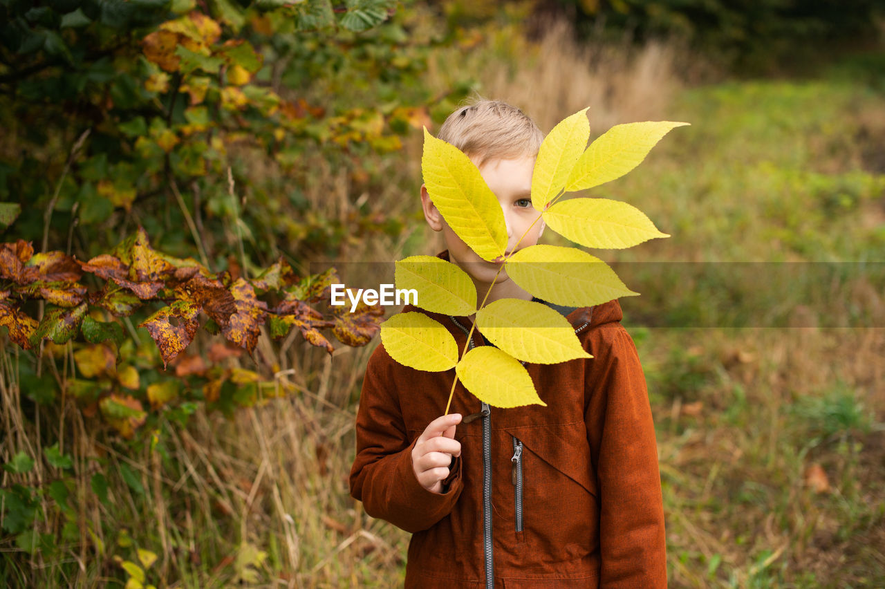 A boy stands on the street and holds a yellow branch with leaves in front of his face