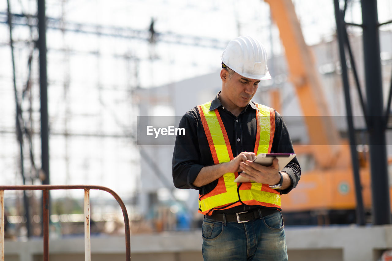 MIDSECTION OF MAN USING MOBILE PHONE WHILE STANDING ON CONSTRUCTION SITE