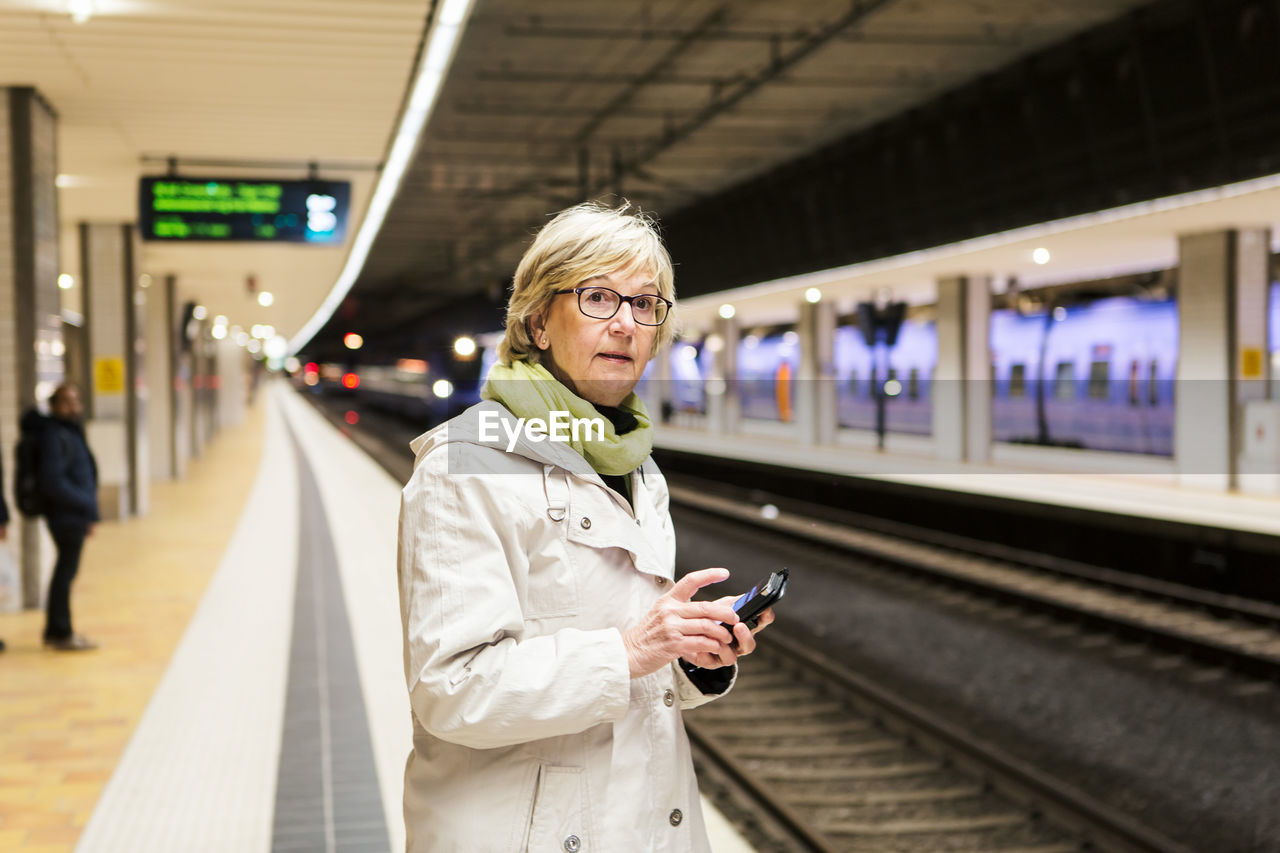 Senior woman looking away while standing with smart phone at subway station