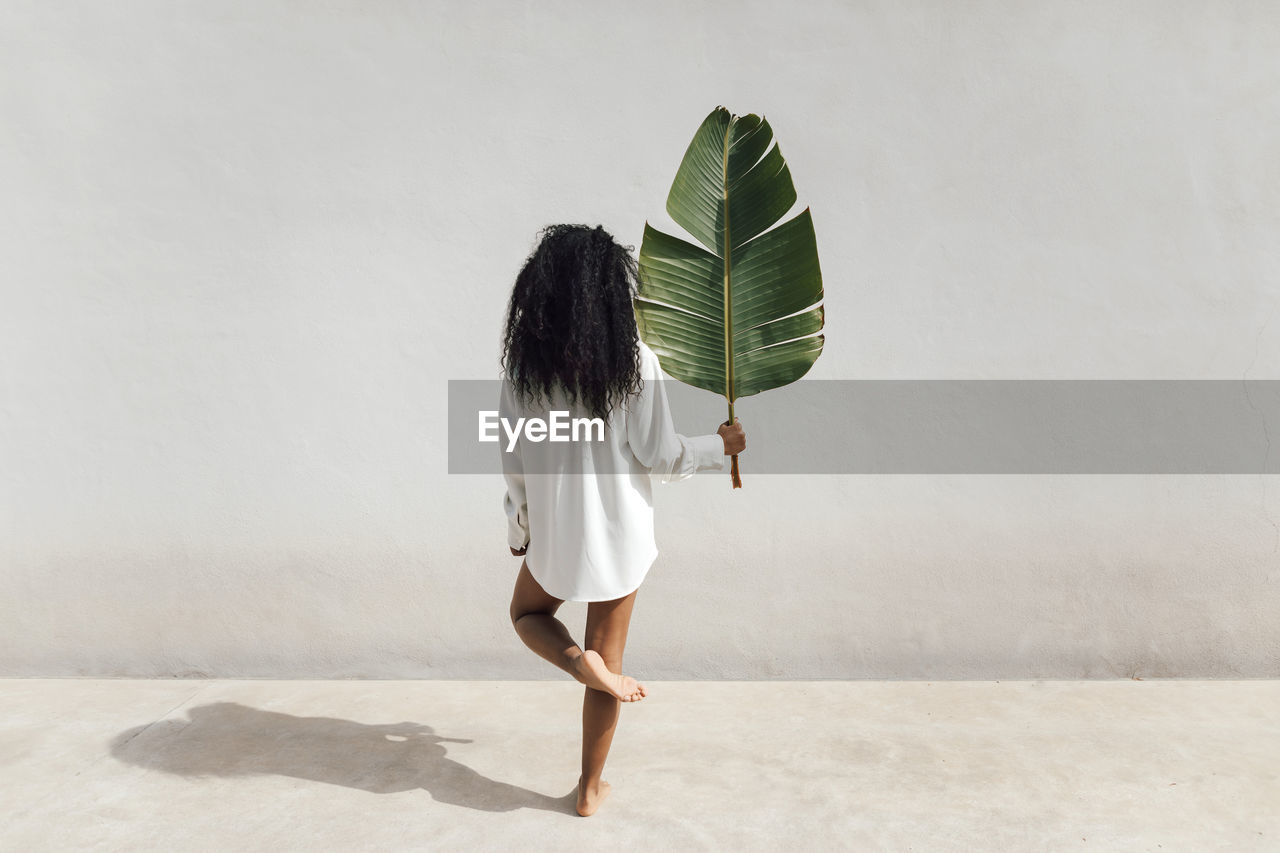 Young woman holding banana leaf while standing on one leg in front of white wall during sunny day