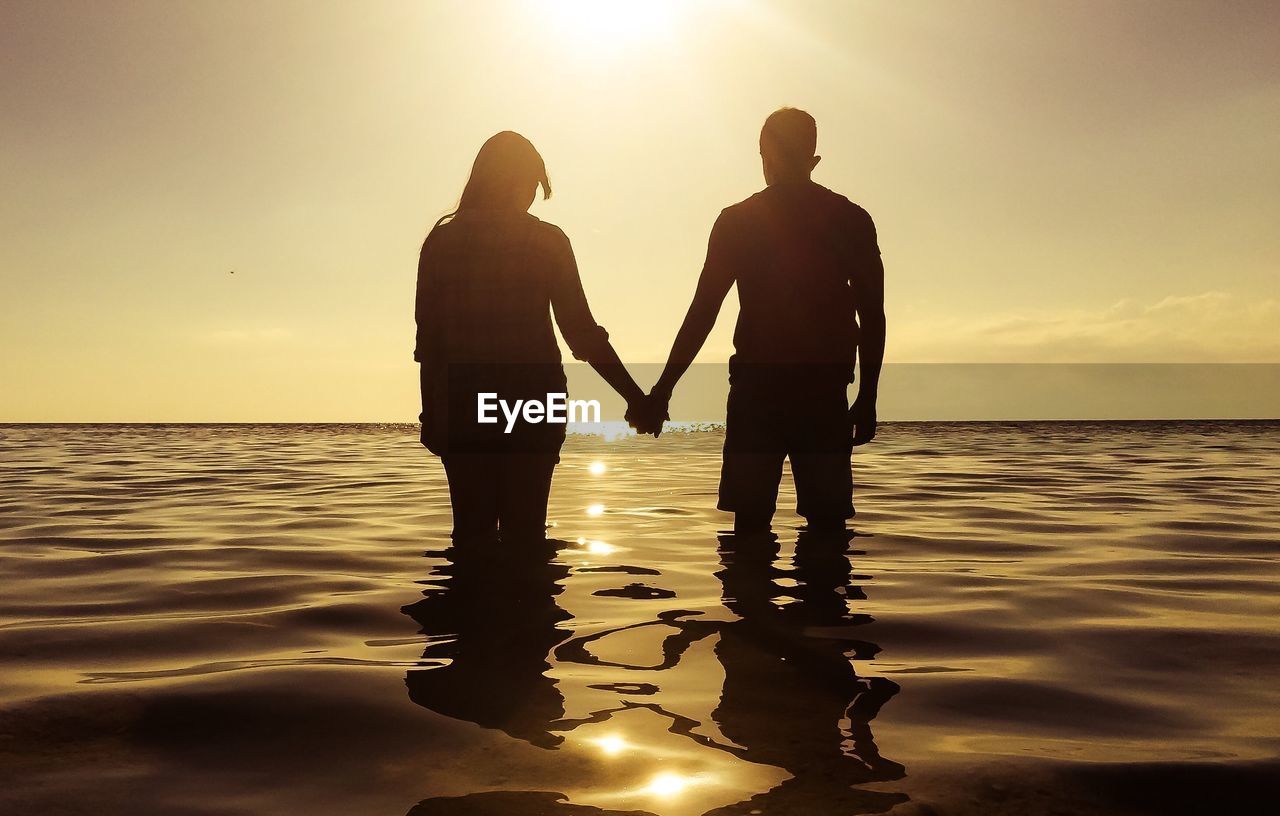 Silhouette couple holding hands while standing in sea against sky during sunset