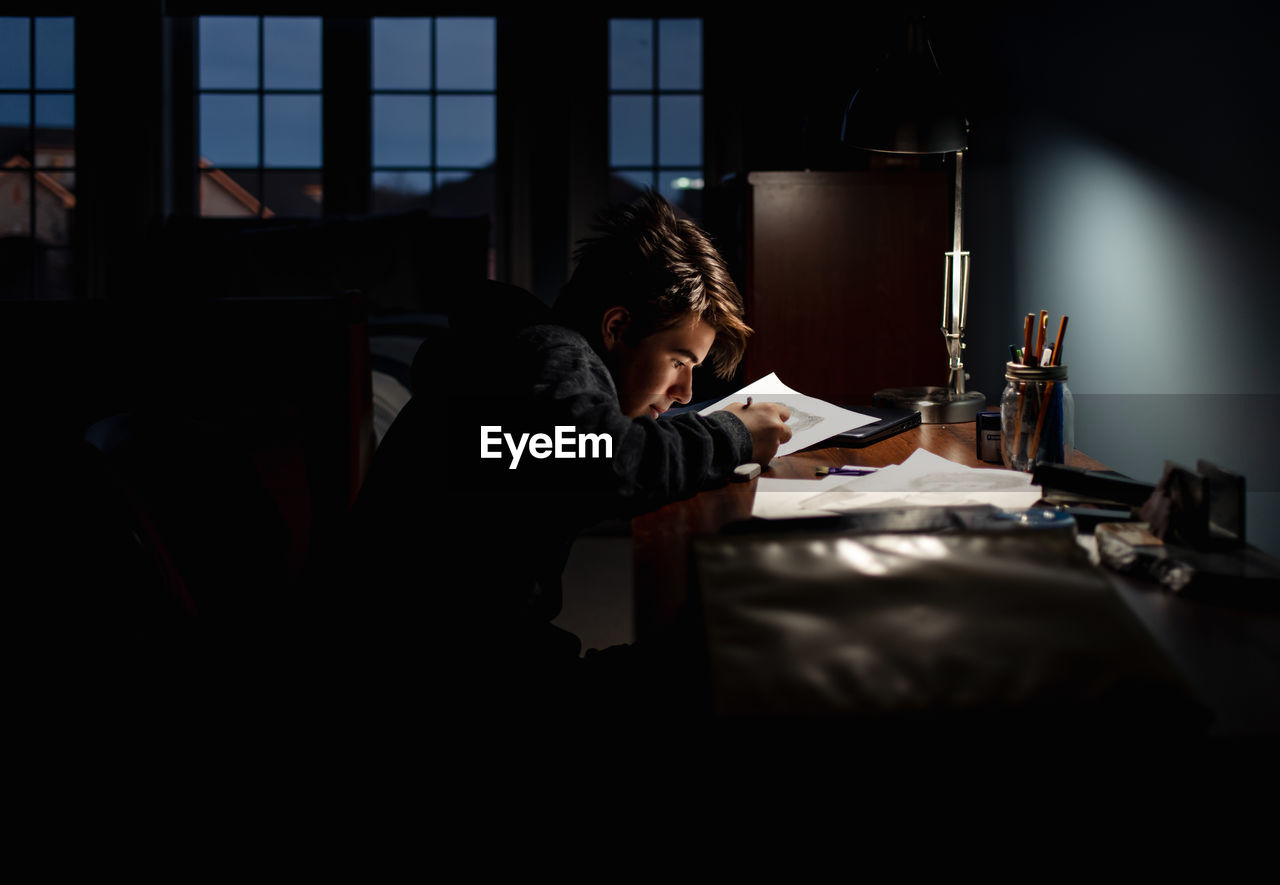 Teenage boy drawing at a desk in a dark room by lamp light.