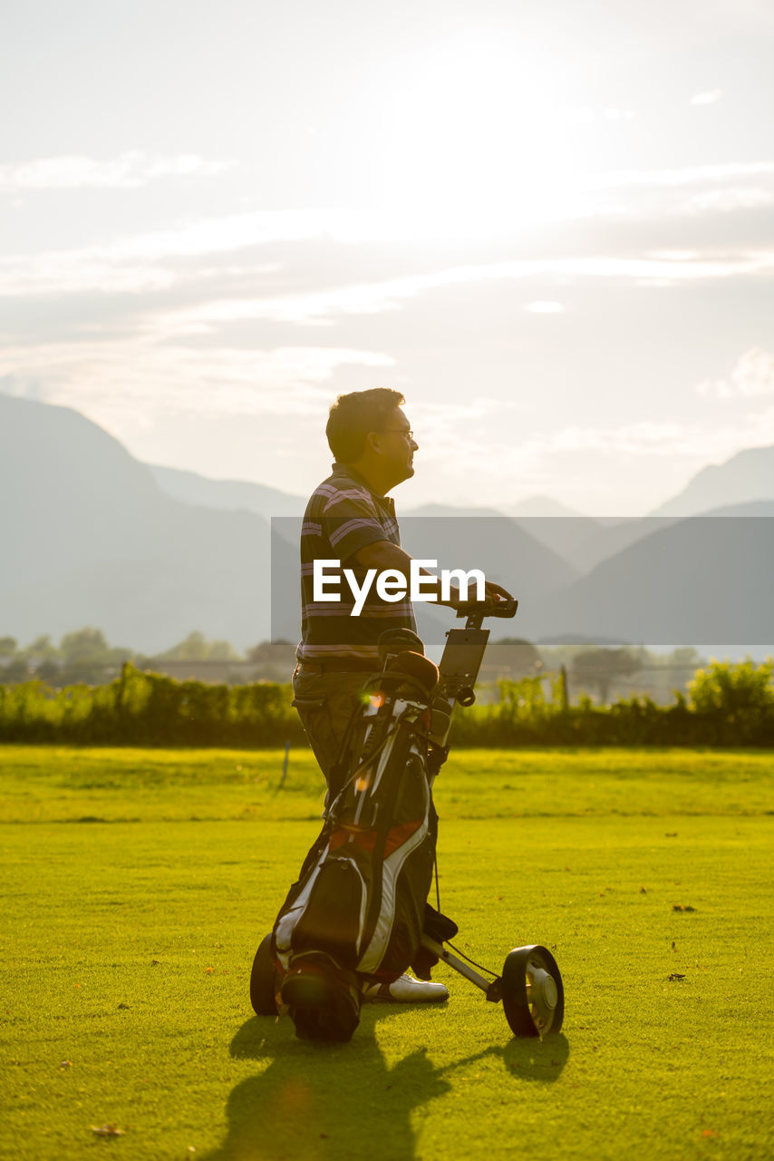 Man standing with golf bag against sky during sunset