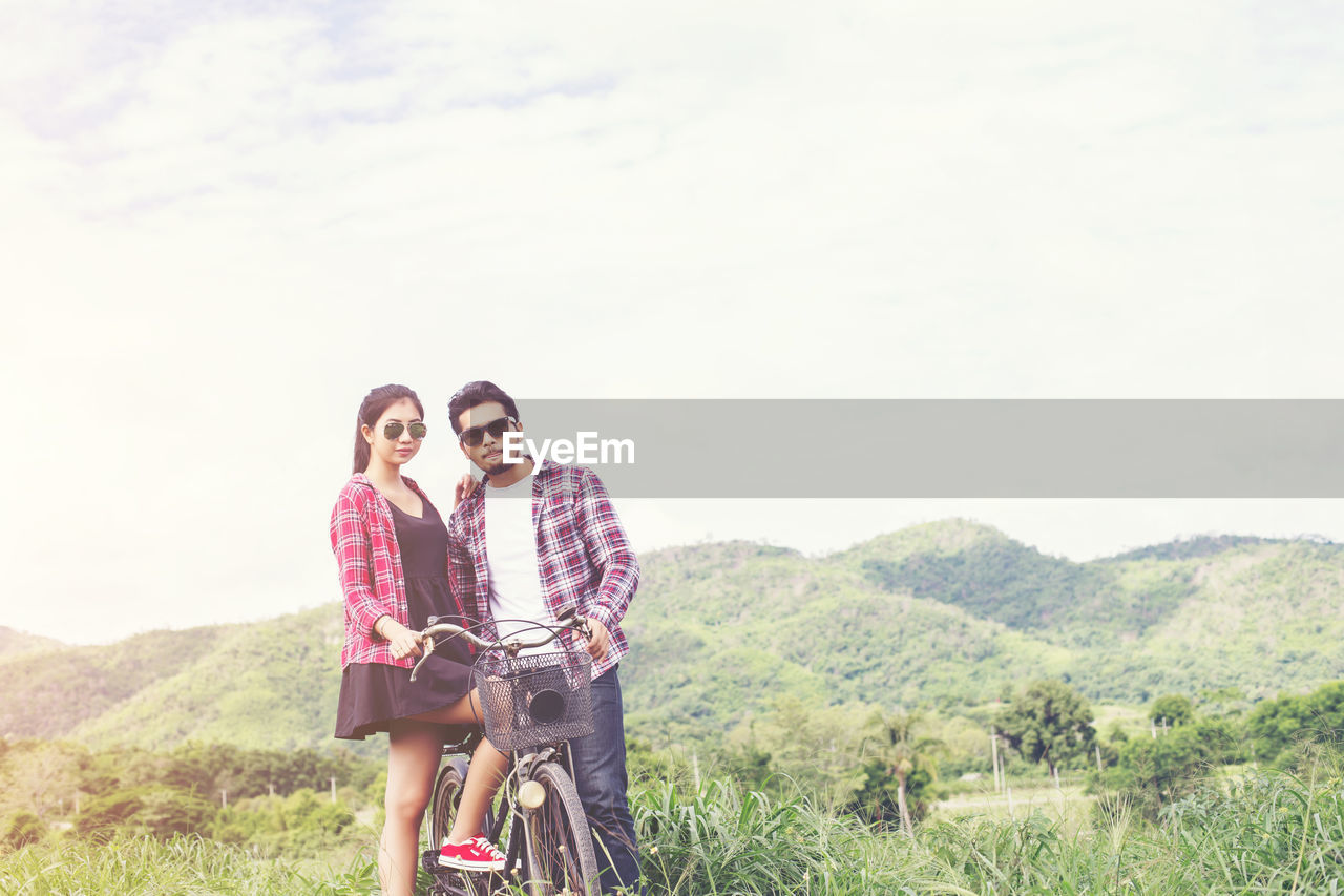 Young couple embracing while standing by bicycle against cloudy sky
