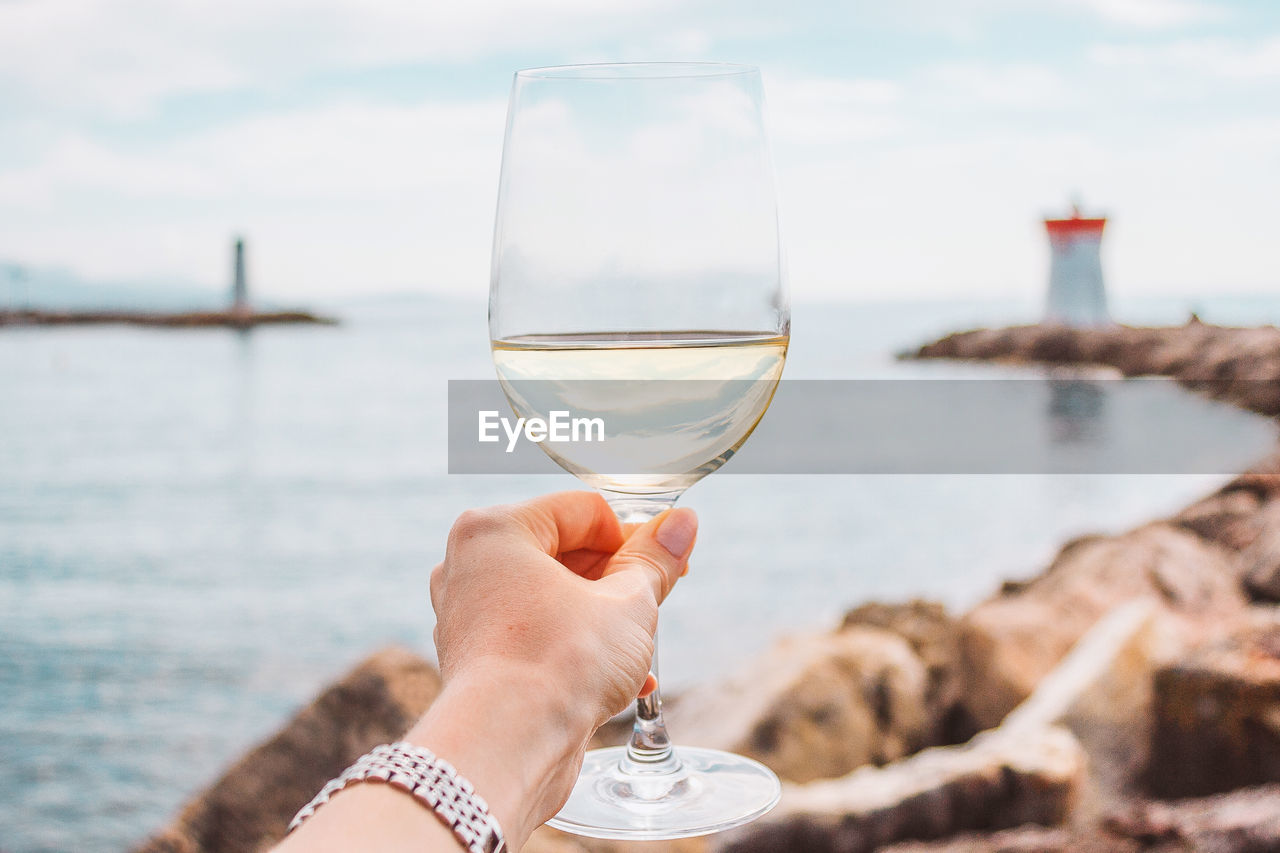 Woman hand with white wine glass on a sea, stones and lighthouse background. nice, french riviera