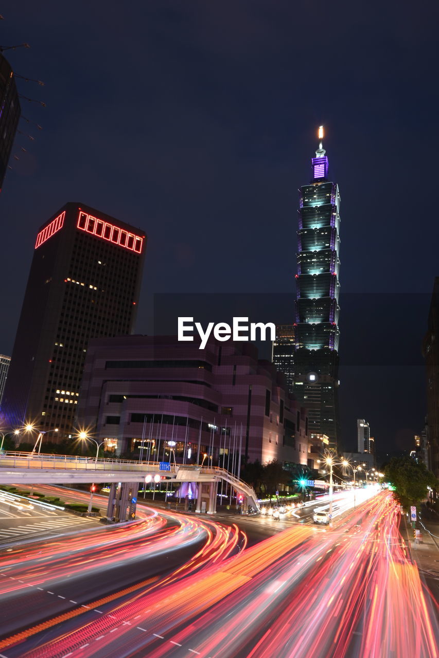 LIGHT TRAILS ON ROAD BY BUILDINGS AGAINST SKY AT NIGHT