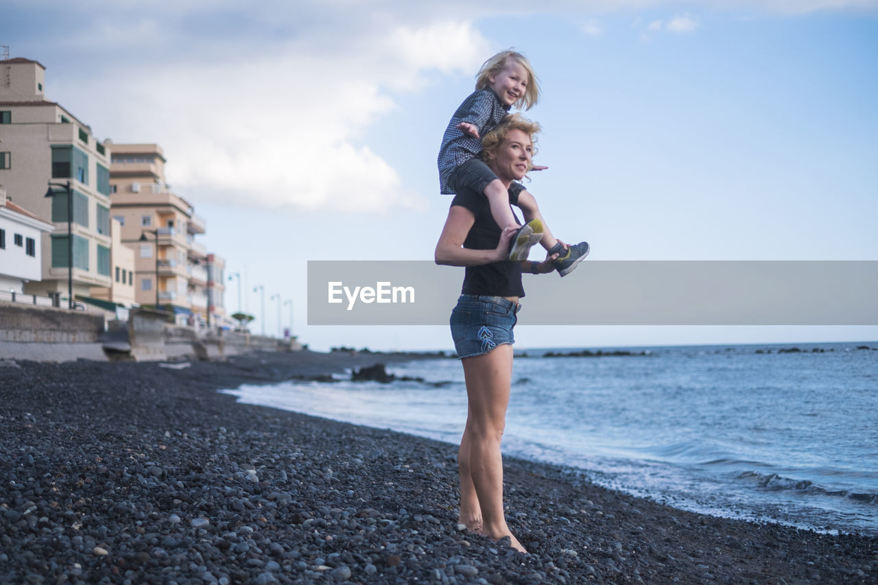 Full length of mother carrying cute son on shoulders while standing at beach against sky during sunset