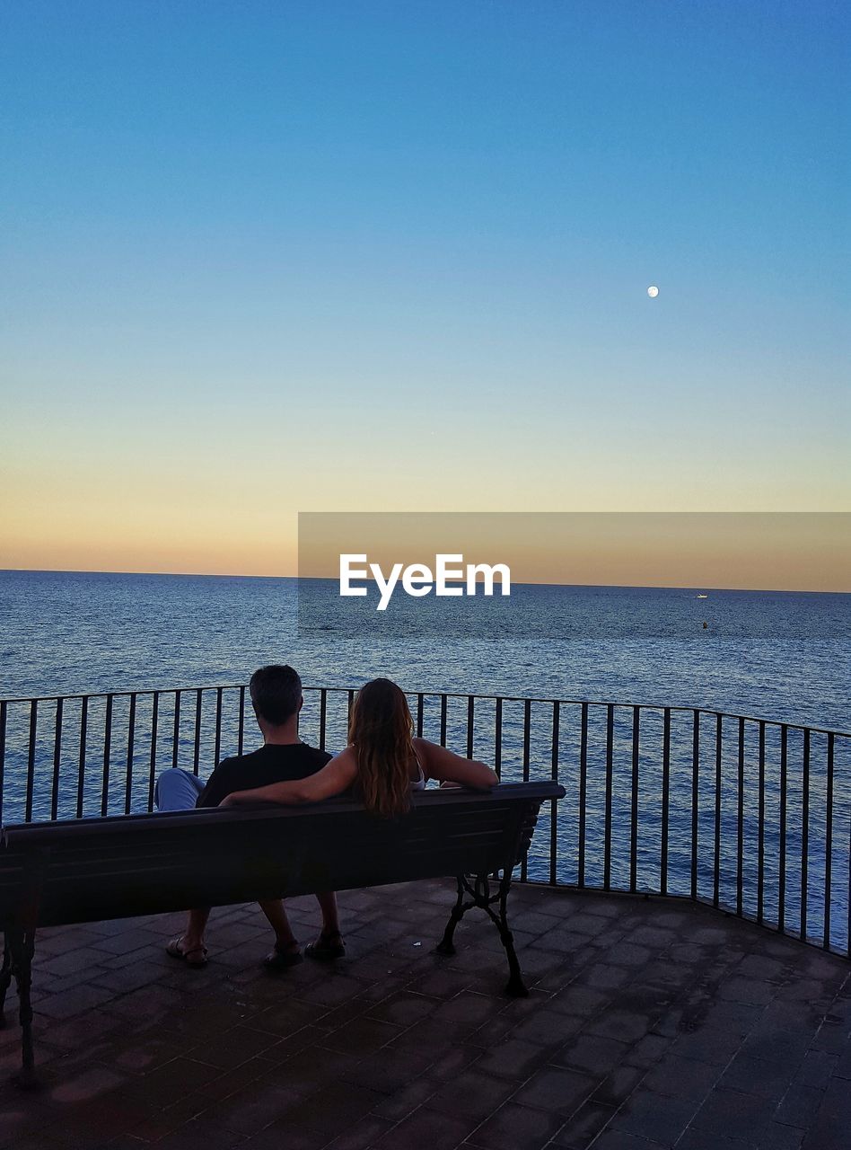 Rear view of couple sitting on bench by railing over sea against sky during sunset