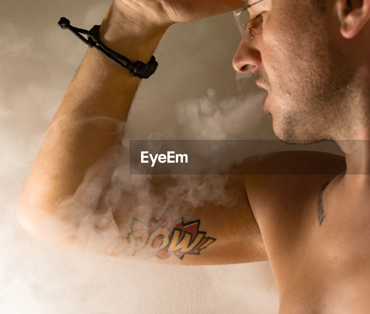 Close-up of shirtless man exhaling smoke against wall