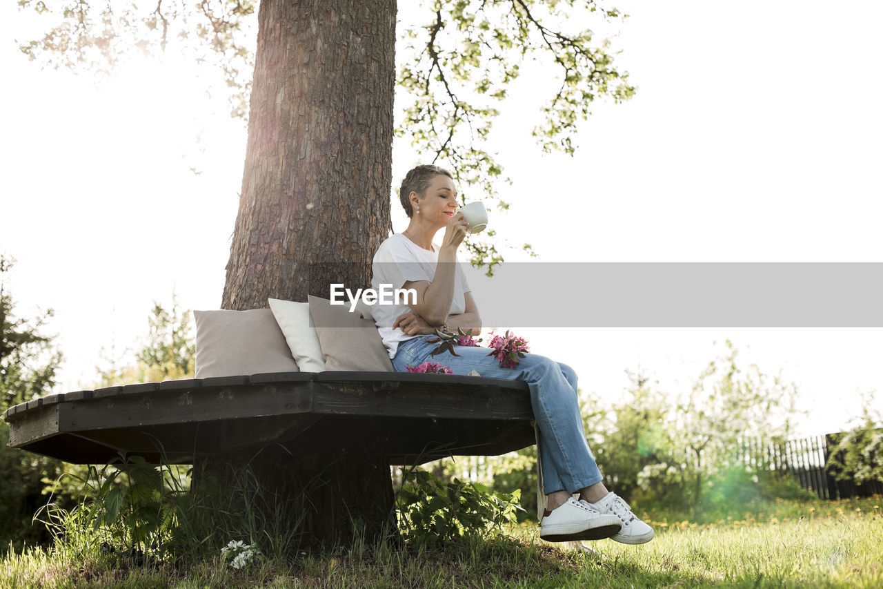 Smiling woman holding coffee cup sitting on bench around tree trunk in garden